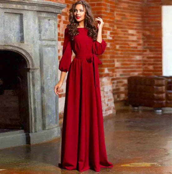 Wine Red - A woman posing in a floor-length white crepe maxi dress featuring a boat neckline, three-quarter balloon sleeves, and a self-tie fabric sash at the waist, set against a modern interior backdrop.