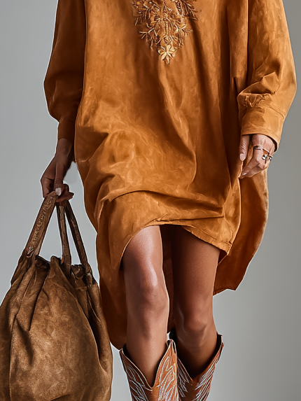 Brown - A model wearing a long-sleeved, ochre-coloured matte satin shift dress featuring a deep V-neckline with intricate floral embroidery, styled with knee-high tan cowboy boots.