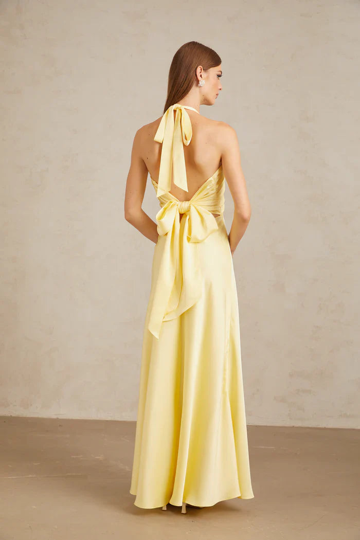 A woman wearing a pale yellow, formal halter neck satin maxi dress with a ruched bust and flowing silhouette against a neutral background.