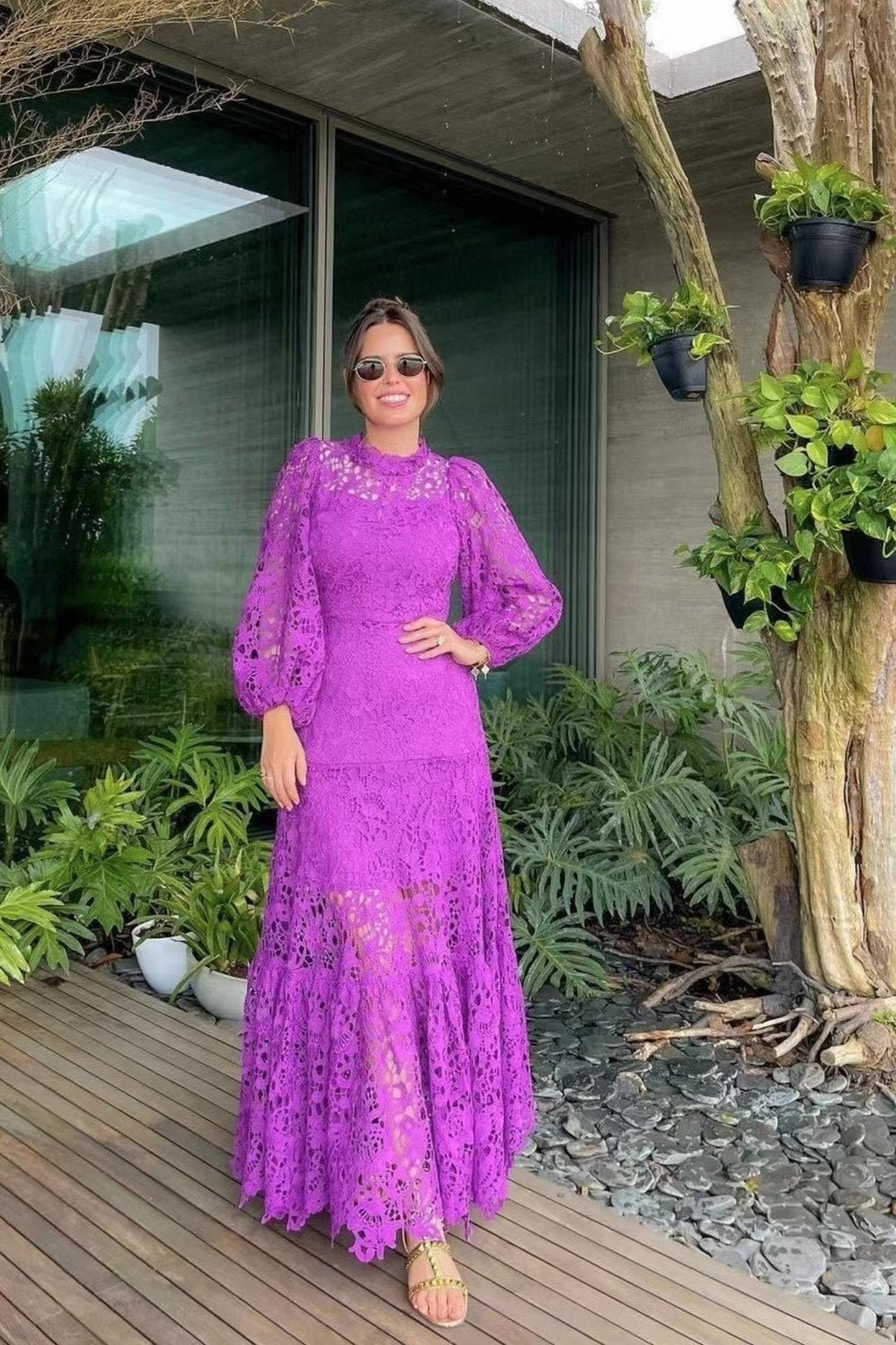 Purple - A woman models a vibrant green, long-sleeve maxi dress featuring an intricate floral lace overlay, a high neckline, and a tiered skirt, holding a matching green clutch in a greenhouse setting.