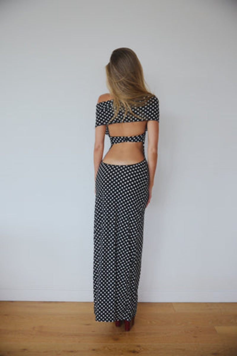 Black - A woman wearing a black and white polka dot maxi dress with halter straps and draped off-the-shoulder sleeves, posing against a white wall.