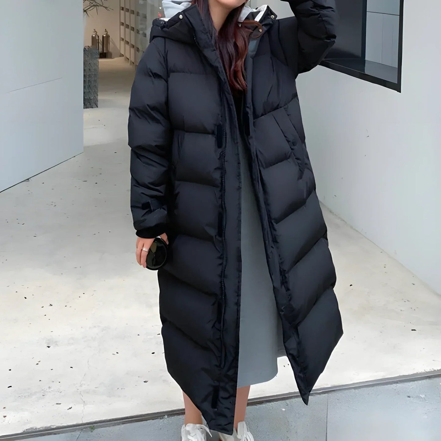 White - A woman walking while wearing a longline, matte black quilted puffer coat with a hood, styled over a light grey outfit and chunky white sneakers.