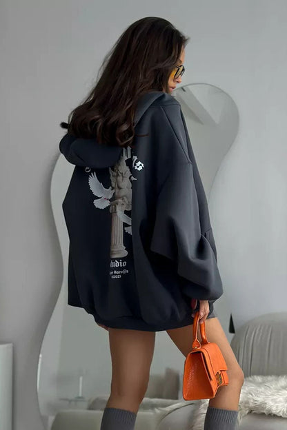 Black - Back view of a woman wearing a dark grey oversized hoodie featuring a large graphic print of an angel statue and white doves, paired with a dark grey pleated mini skirt.