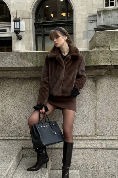 A woman stands outdoors in an urban setting wearing a plush, dark brown cropped faux fur jacket with a wide collar, styled with a mini skirt and knee-high boots.