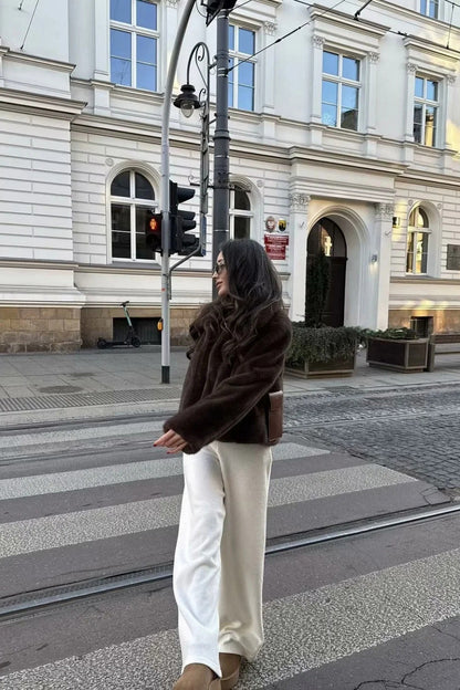 A woman stands outdoors in an urban setting wearing a plush, dark brown cropped faux fur jacket with a wide collar, styled with a mini skirt and knee-high boots.