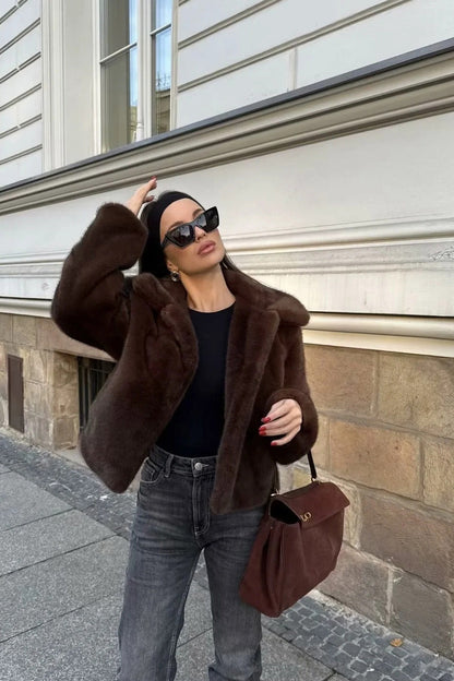 A woman stands outdoors in an urban setting wearing a plush, dark brown cropped faux fur jacket with a wide collar, styled with a mini skirt and knee-high boots.