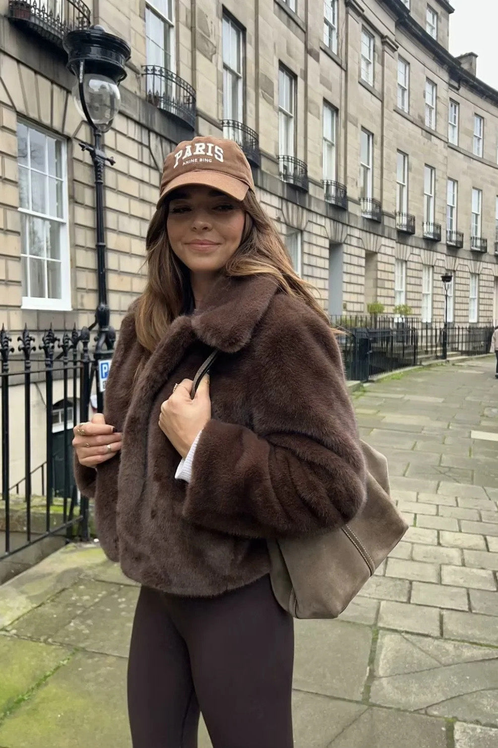 A woman stands outdoors in an urban setting wearing a plush, dark brown cropped faux fur jacket with a wide collar, styled with a mini skirt and knee-high boots.