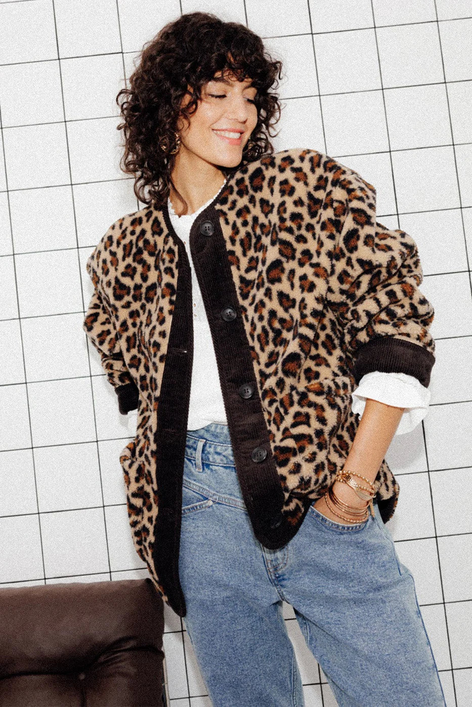A woman wearing a relaxed-fit, fuzzy knit cardigan featuring a bold leopard print and contrasting dark ribbed placket, styled open over a white top and blue jeans.
