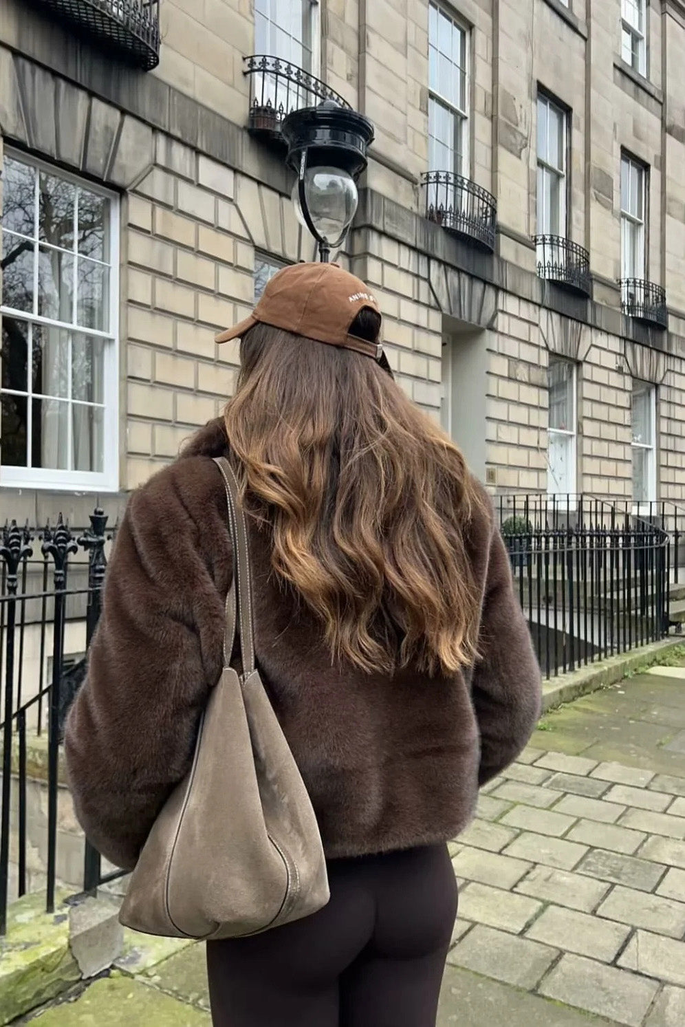A woman stands outdoors in an urban setting wearing a plush, dark brown cropped faux fur jacket with a wide collar, styled with a mini skirt and knee-high boots.