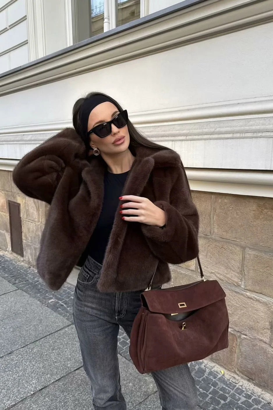 Brown - A woman stands outdoors in an urban setting wearing a plush, dark brown cropped faux fur jacket with a wide collar, styled with a mini skirt and knee-high boots.