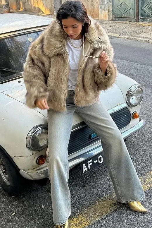 Brown - A woman sits on the hood of a vintage car wearing a fluffy, light brown faux fur cropped jacket with a large collar, styled with light wash wide-leg jeans.