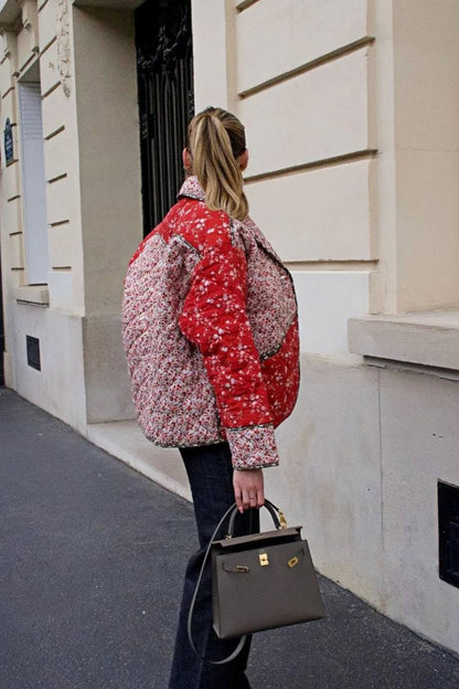 A woman wearing a hip-length quilted jacket featuring a mixed red and white floral print, contrast dark piping, and a button-front closure.