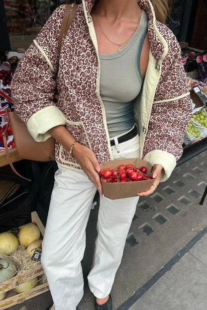 A woman wearing a quilted, floral print jacket with contrast piping and rolled cuffs over a ribbed tank top and white trousers.