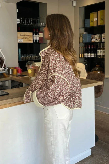 A woman wearing a quilted, floral print jacket with contrast piping and rolled cuffs over a ribbed tank top and white trousers.