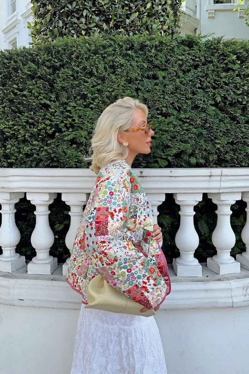 A woman wearing a vibrant, multi-floral patchwork quilted jacket with a collarless neckline and contrast piping, paired with a white skirt.