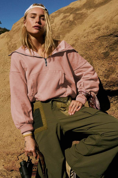 A woman wearing a dusty pink oversized half-zip sweatshirt with a high funnel neck, kangaroo pocket, and ribbed cuffs, paired with distressed denim shorts.