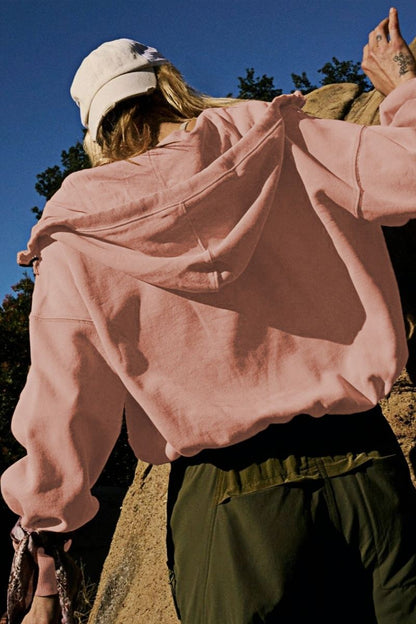 A woman wearing a dusty pink oversized half-zip sweatshirt with a high funnel neck, kangaroo pocket, and ribbed cuffs, paired with distressed denim shorts.