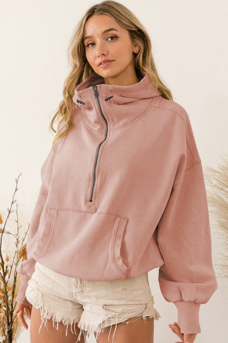 A woman wearing a dusty pink oversized half-zip sweatshirt with a high funnel neck, kangaroo pocket, and ribbed cuffs, paired with distressed denim shorts.