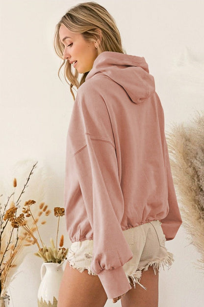 A woman wearing a dusty pink oversized half-zip sweatshirt with a high funnel neck, kangaroo pocket, and ribbed cuffs, paired with distressed denim shorts.