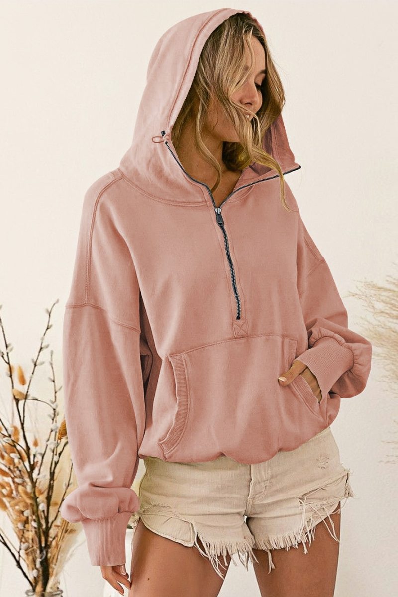A woman wearing a dusty pink oversized half-zip sweatshirt with a high funnel neck, kangaroo pocket, and ribbed cuffs, paired with distressed denim shorts.