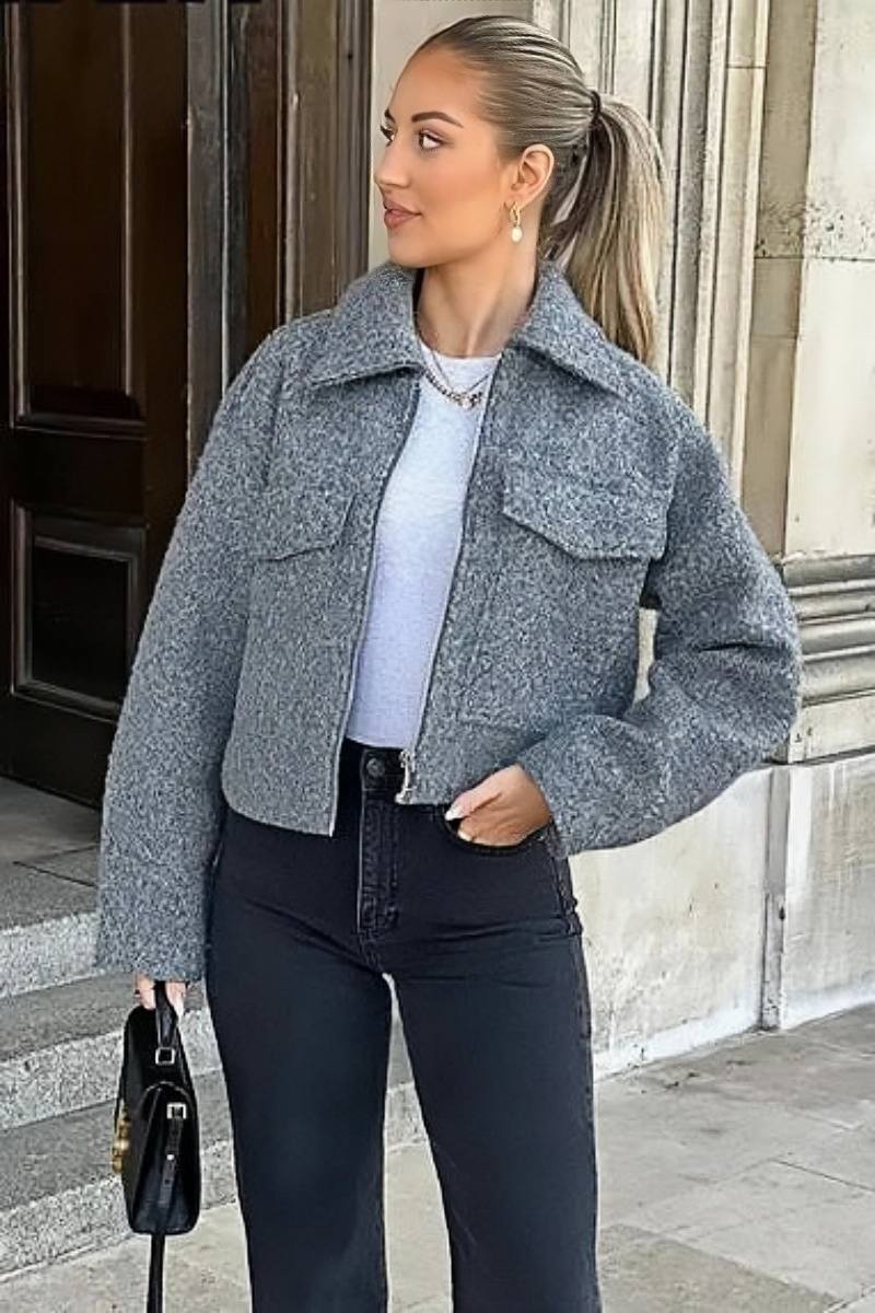 A woman wearing a women's grey textured cropped jacket with a silver front zipper and chest flap pockets, styled over a white t-shirt and black pleated mini skirt.
