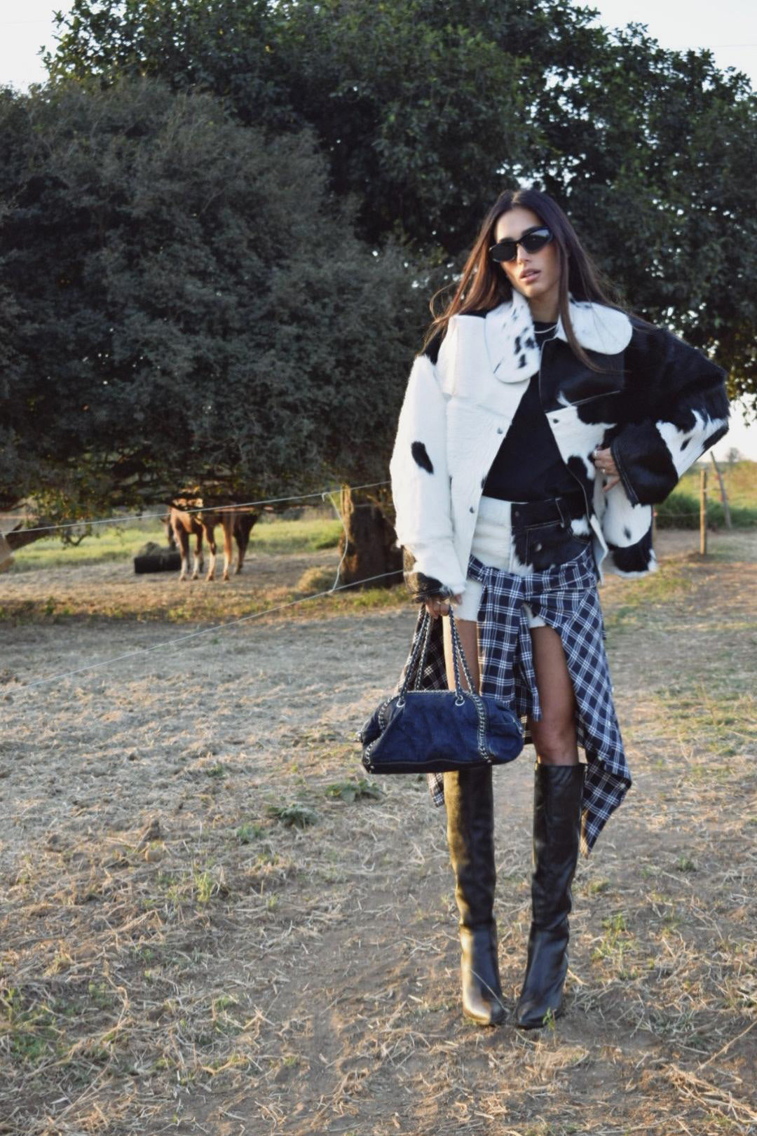 Woman standing outdoors wearing an oversized black and white cow print faux fur jacket, styled with a plaid shirt tied around her waist and tall black boots.