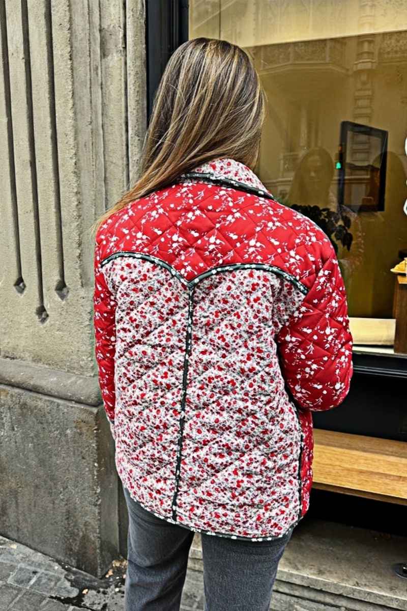A woman wearing a hip-length quilted jacket featuring a mixed red and white floral print, contrast dark piping, and a button-front closure.