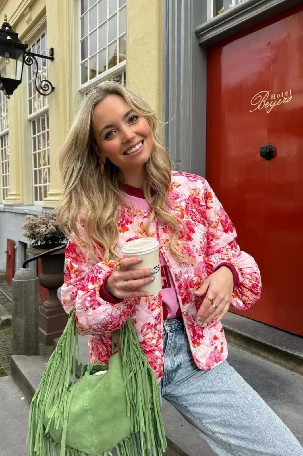 Red - Woman wearing a vibrant pink and red floral quilted bomber jacket with snap buttons, paired with light wash denim jeans and carrying a green fringed bag.