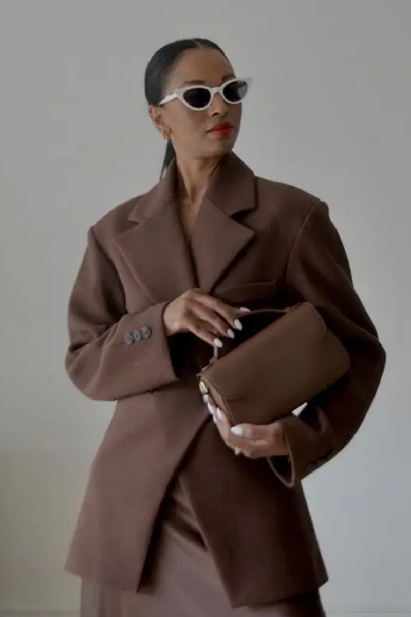 Brown - A woman wearing a brown oversized double-breasted blazer with peak lapels over a matching satin midi skirt.