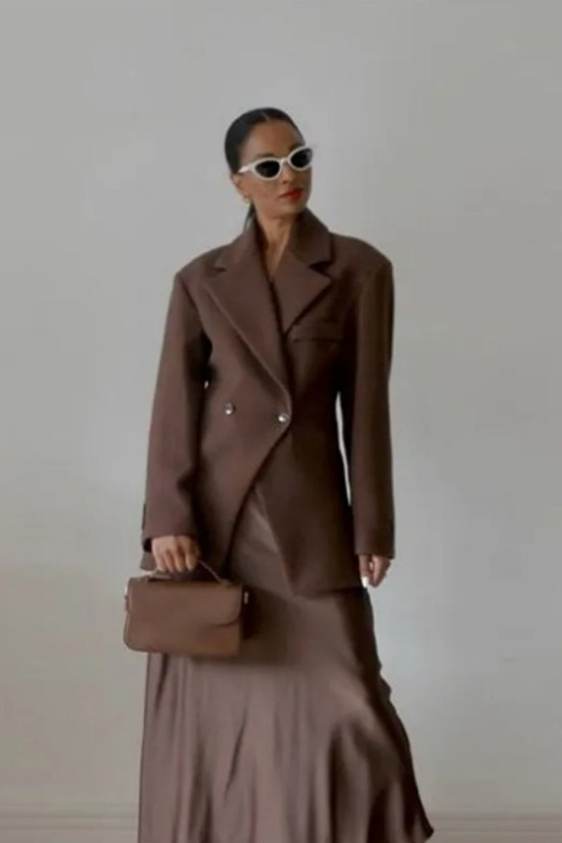 Brown - A woman wearing a brown oversized double-breasted blazer with peak lapels over a matching satin midi skirt.