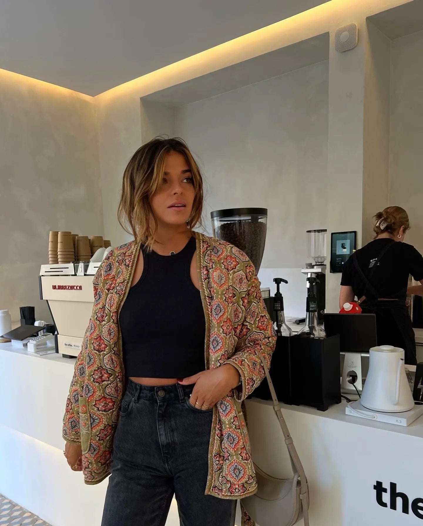 Multicolor - Woman in a cafe wearing an open-front quilted jacket with a colourful geometric medallion print over a black tank top and dark denim jeans.