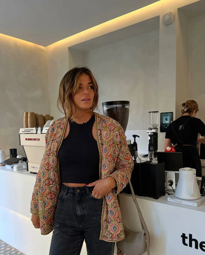 Multicolor - Woman in a cafe wearing an open-front quilted jacket with a colourful geometric medallion print over a black tank top and dark denim jeans.