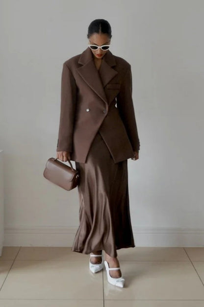 Brown - A woman wearing a brown oversized double-breasted blazer with peak lapels over a matching satin midi skirt.