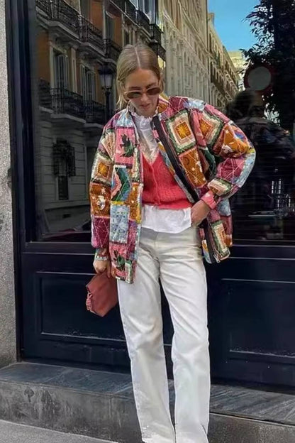 A woman wearing a vibrant, multi-coloured quilted patchwork jacket over a pink cardigan and white shirt, paired with white trousers.