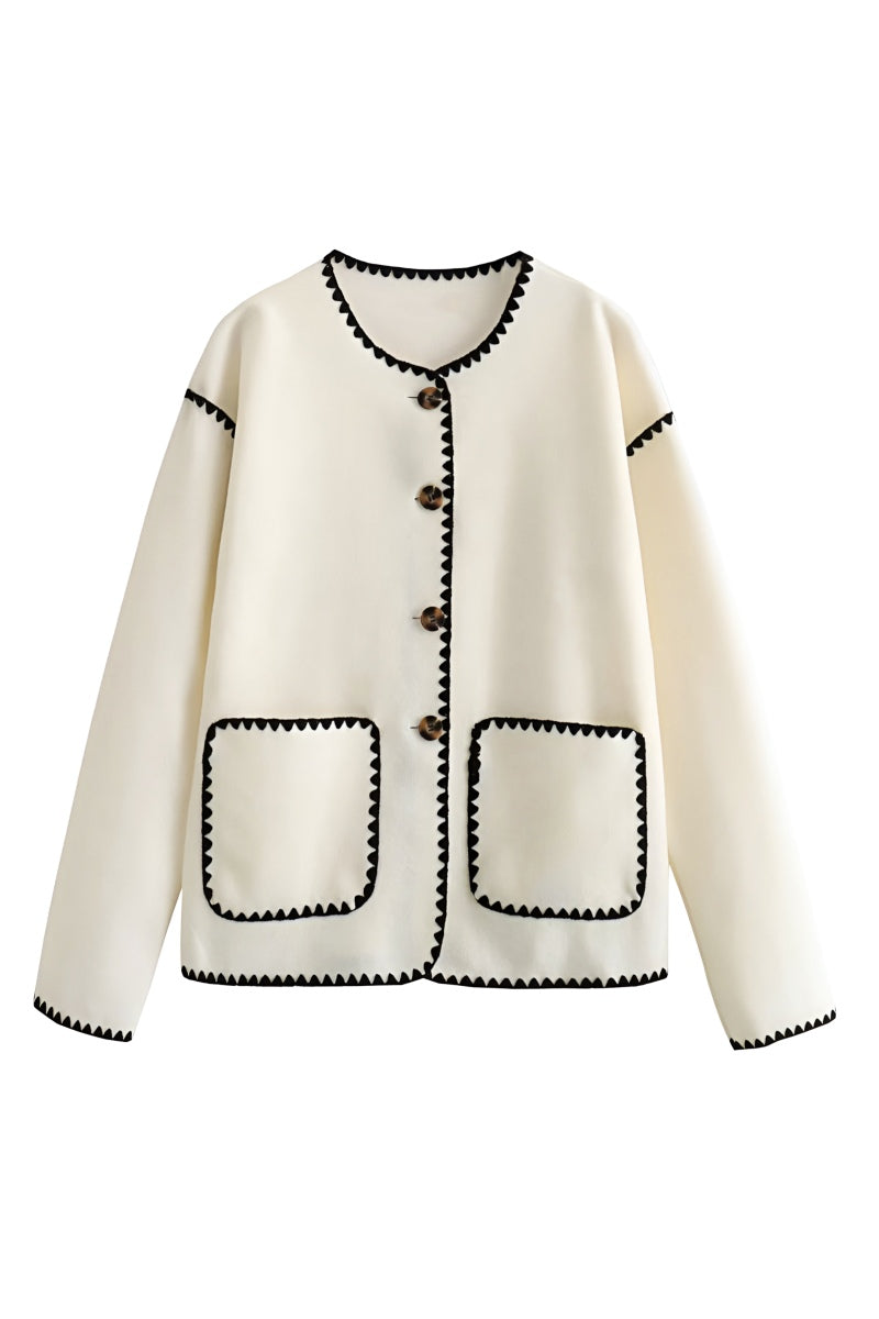 A woman wearing a cream-coloured knit cardigan featuring black contrast stitching along the edges, neckline, and front patch pockets, styled with dark denim jeans.