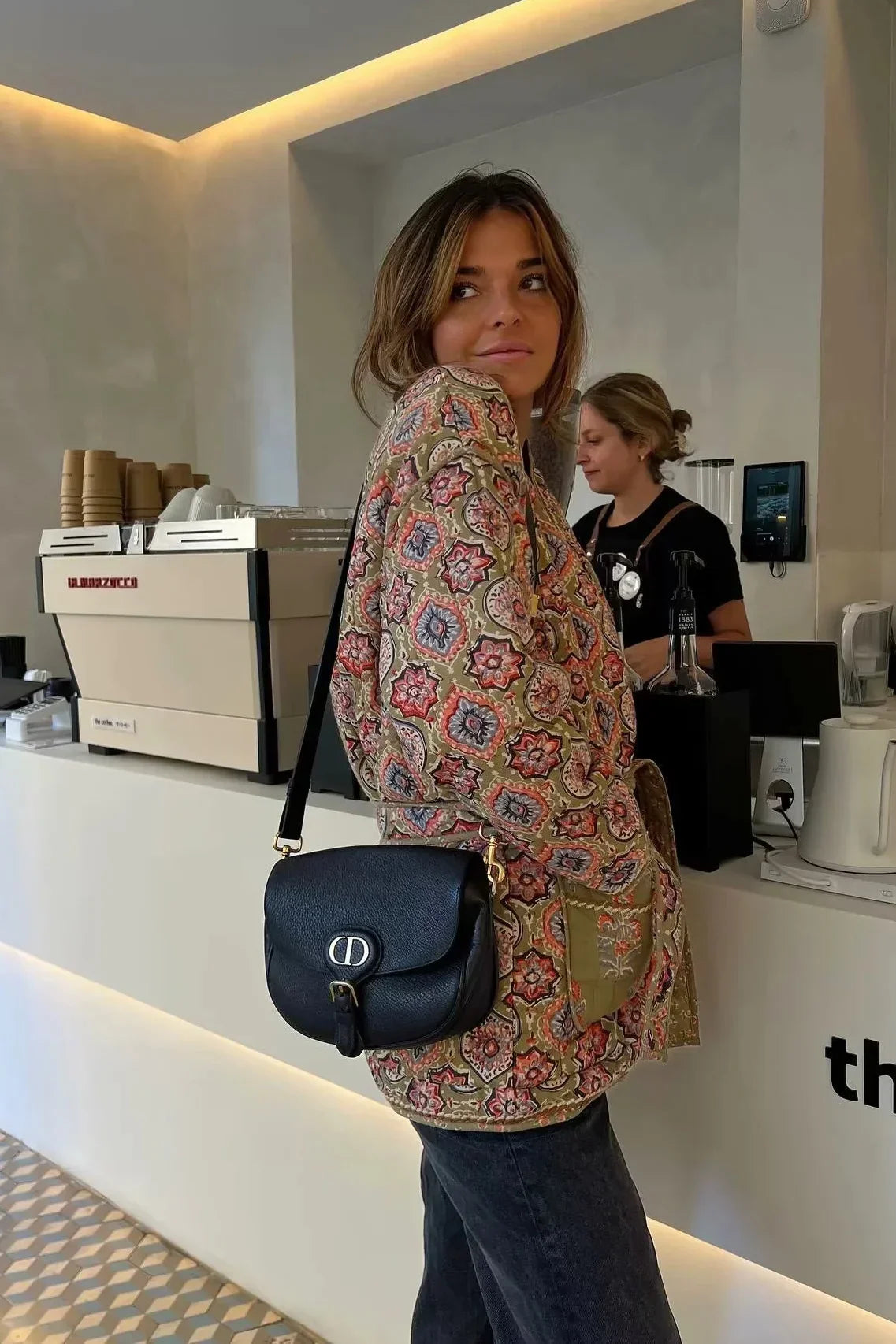 Multicolor - Woman in a cafe wearing an open-front quilted jacket with a colourful geometric medallion print over a black tank top and dark denim jeans.