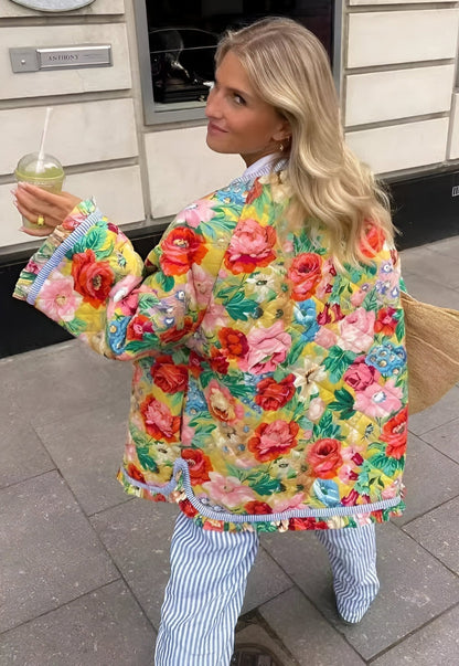 Women's longline quilted jacket featuring a bright, oversized floral print on a yellow background, finished with contrasting blue and white striped trim around the edges and cuffs, and a wooden-style button-front closure.