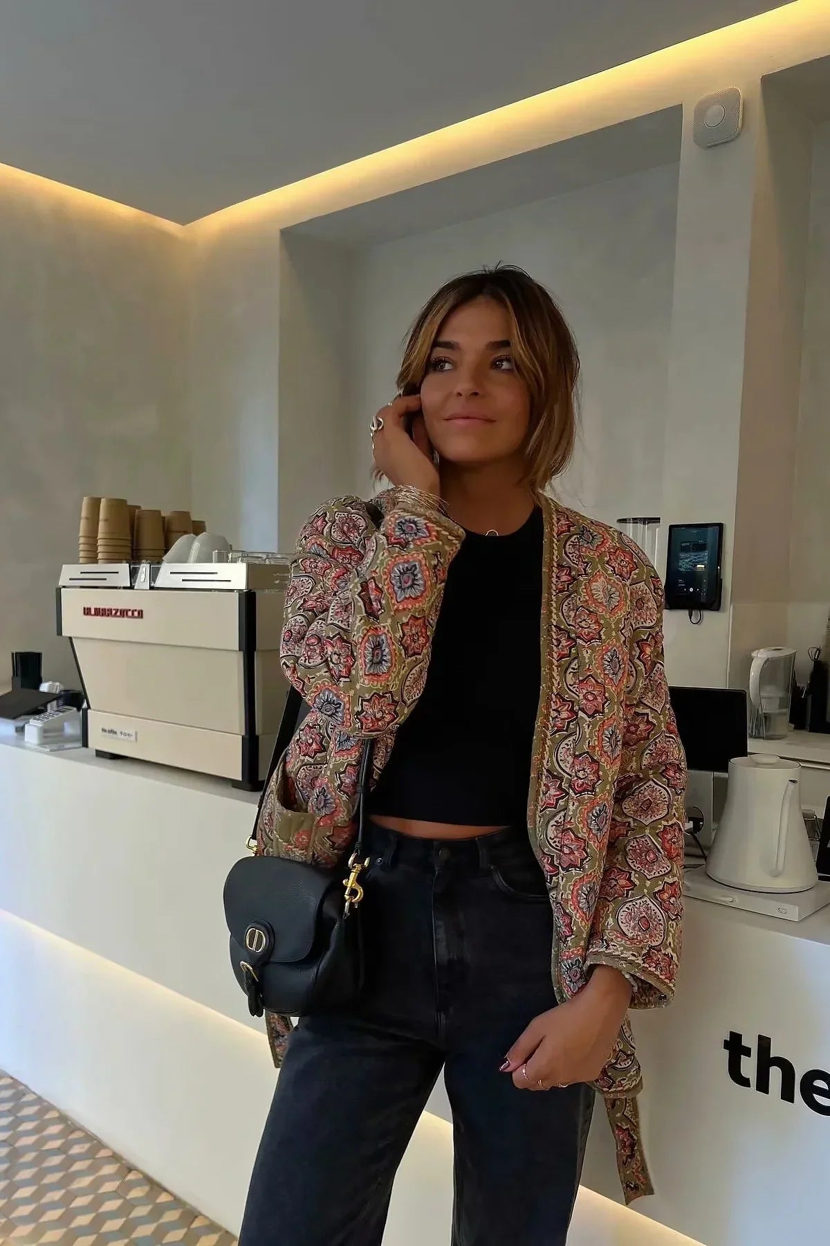 Multicolor - Woman in a cafe wearing an open-front quilted jacket with a colourful geometric medallion print over a black tank top and dark denim jeans.