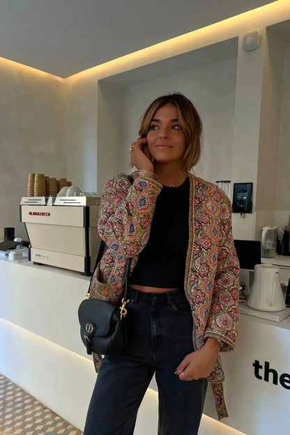 Multicolor - Woman in a cafe wearing an open-front quilted jacket with a colourful geometric medallion print over a black tank top and dark denim jeans.