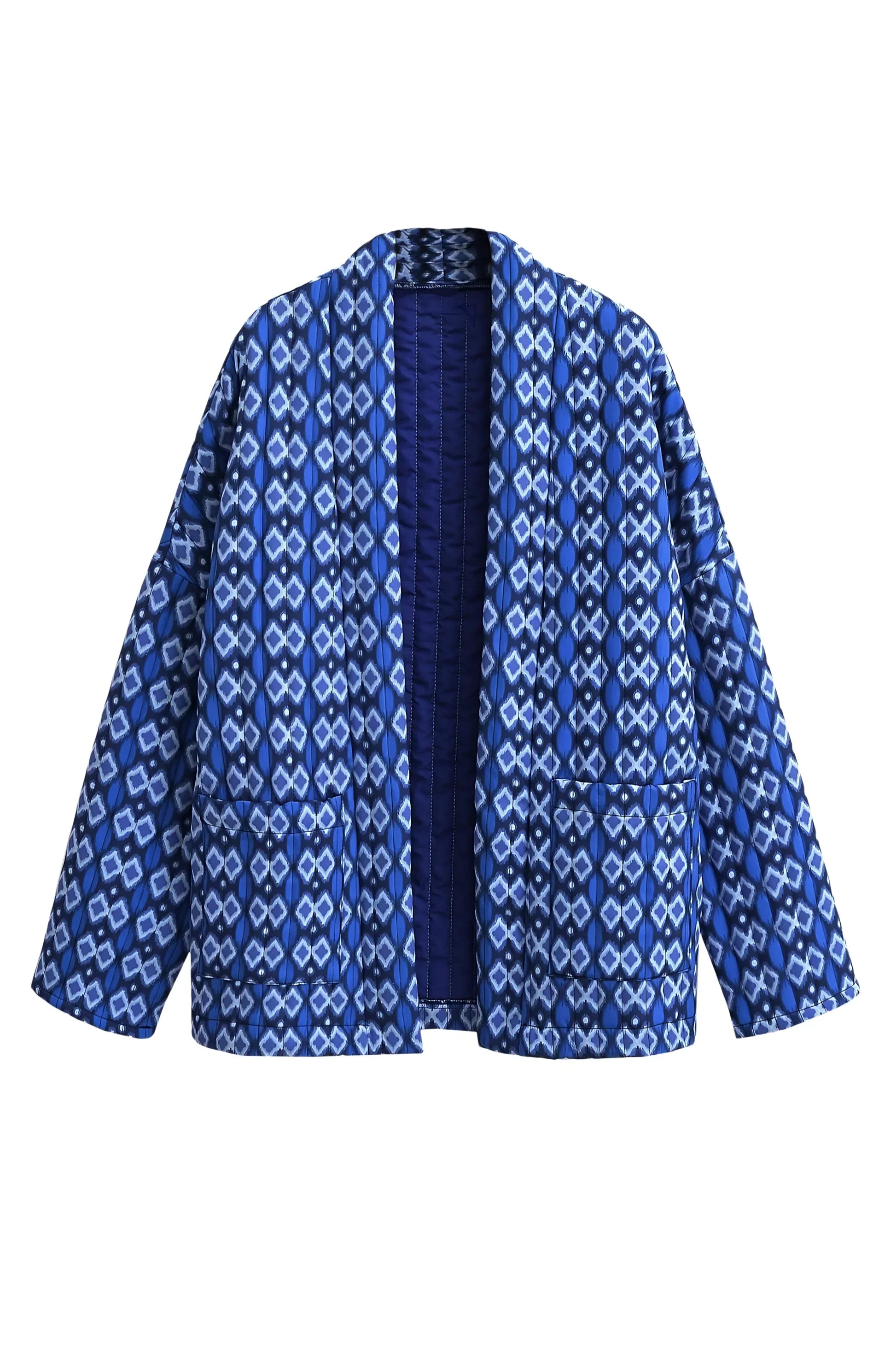 Blue - A woman stands outdoors wearing a relaxed-fit, hip-length quilted jacket featuring a bold blue and white geometric ikat print, styled open over a white crop top and straight-leg jeans.