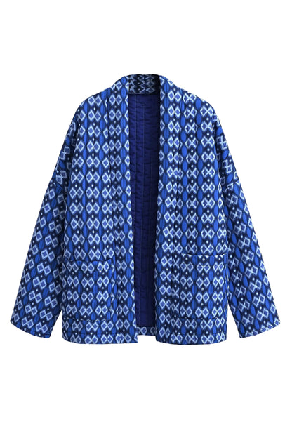 Blue - A woman stands outdoors wearing a relaxed-fit, hip-length quilted jacket featuring a bold blue and white geometric ikat print, styled open over a white crop top and straight-leg jeans.