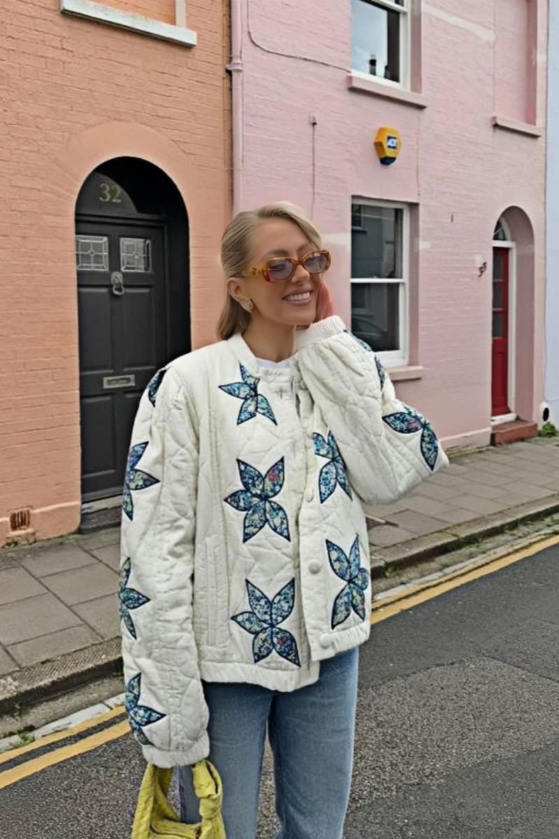 A woman sitting outdoors wearing a cream quilted jacket with blue star appliqués, styled with light wash jeans and sunglasses.