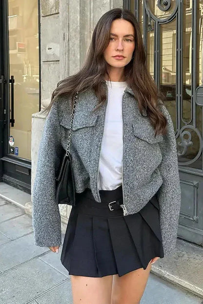 A woman wearing a women's grey textured cropped jacket with a silver front zipper and chest flap pockets, styled over a white t-shirt and black pleated mini skirt.