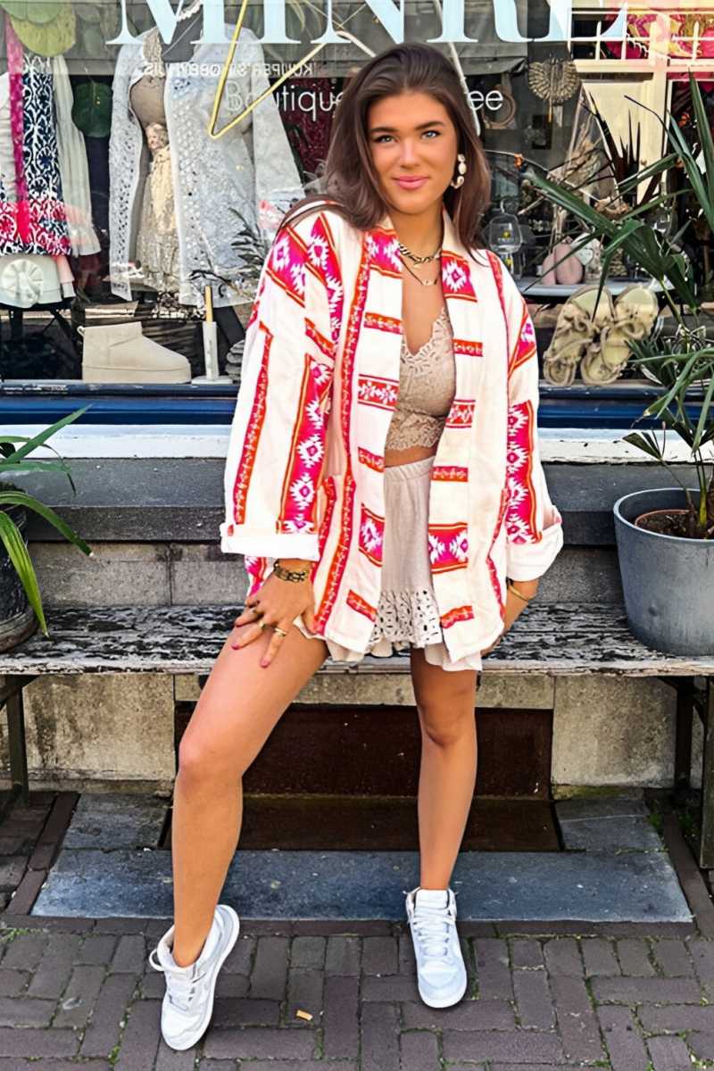 A woman wearing a relaxed, open-front kimono jacket featuring a vibrant pink and orange geometric Aztec print over a cream base, styled over a crochet crop top and shorts.