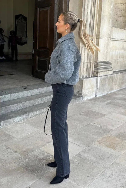A woman wearing a women's grey textured cropped jacket with a silver front zipper and chest flap pockets, styled over a white t-shirt and black pleated mini skirt.