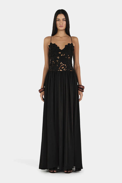 A woman wearing a formal black maxi dress featuring a delicate floral lace bodice with sheer cutouts, thin spaghetti straps, and a long, flowing pleated skirt.
