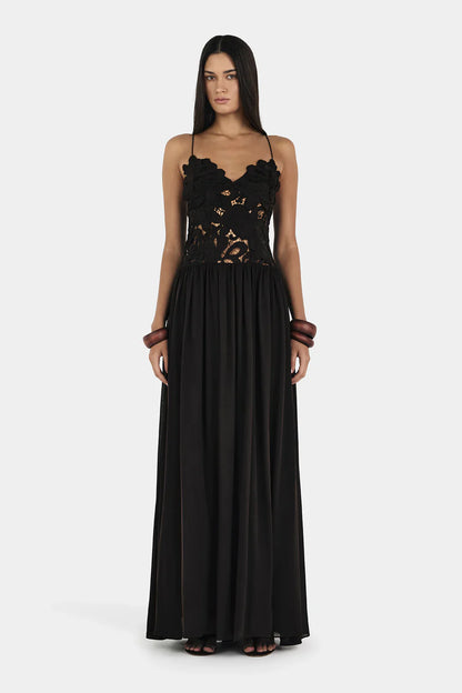 A woman wearing a formal black maxi dress featuring a delicate floral lace bodice with sheer cutouts, thin spaghetti straps, and a long, flowing pleated skirt.