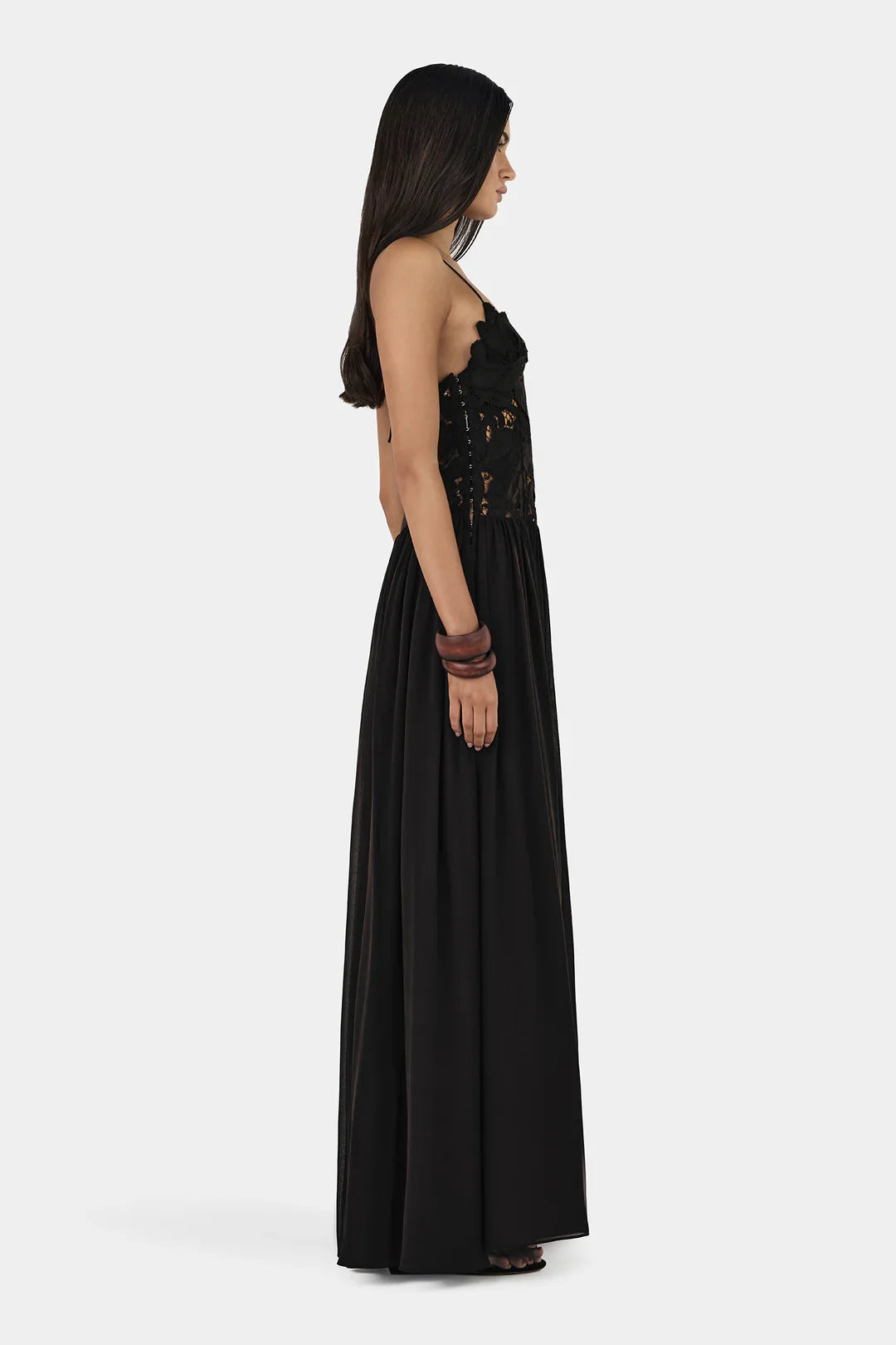A woman wearing a formal black maxi dress featuring a delicate floral lace bodice with sheer cutouts, thin spaghetti straps, and a long, flowing pleated skirt.