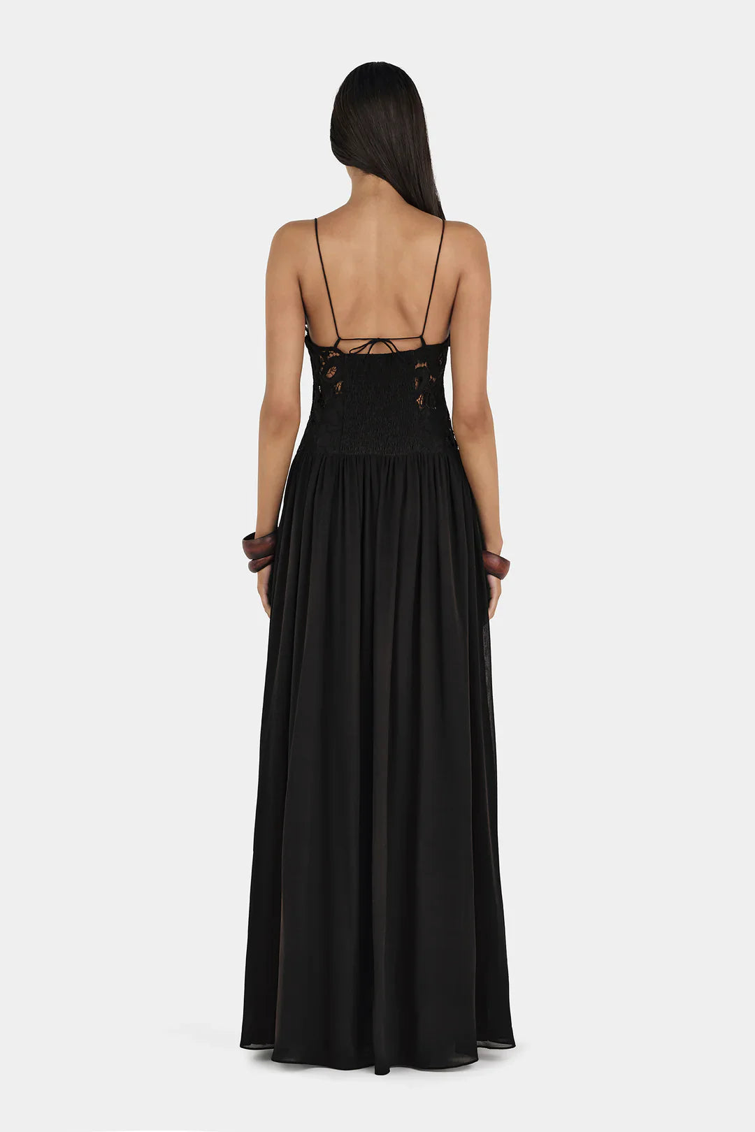 A woman wearing a formal black maxi dress featuring a delicate floral lace bodice with sheer cutouts, thin spaghetti straps, and a long, flowing pleated skirt.