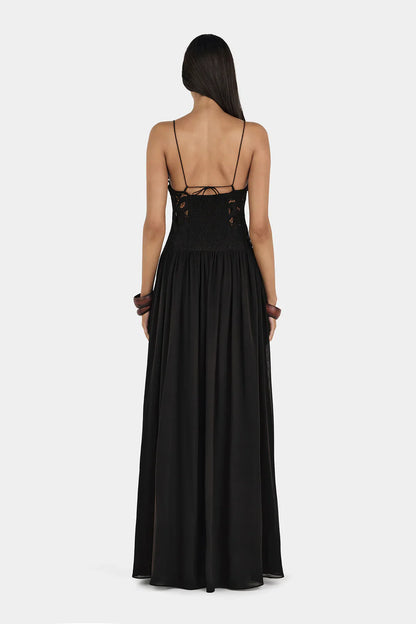 A woman wearing a formal black maxi dress featuring a delicate floral lace bodice with sheer cutouts, thin spaghetti straps, and a long, flowing pleated skirt.